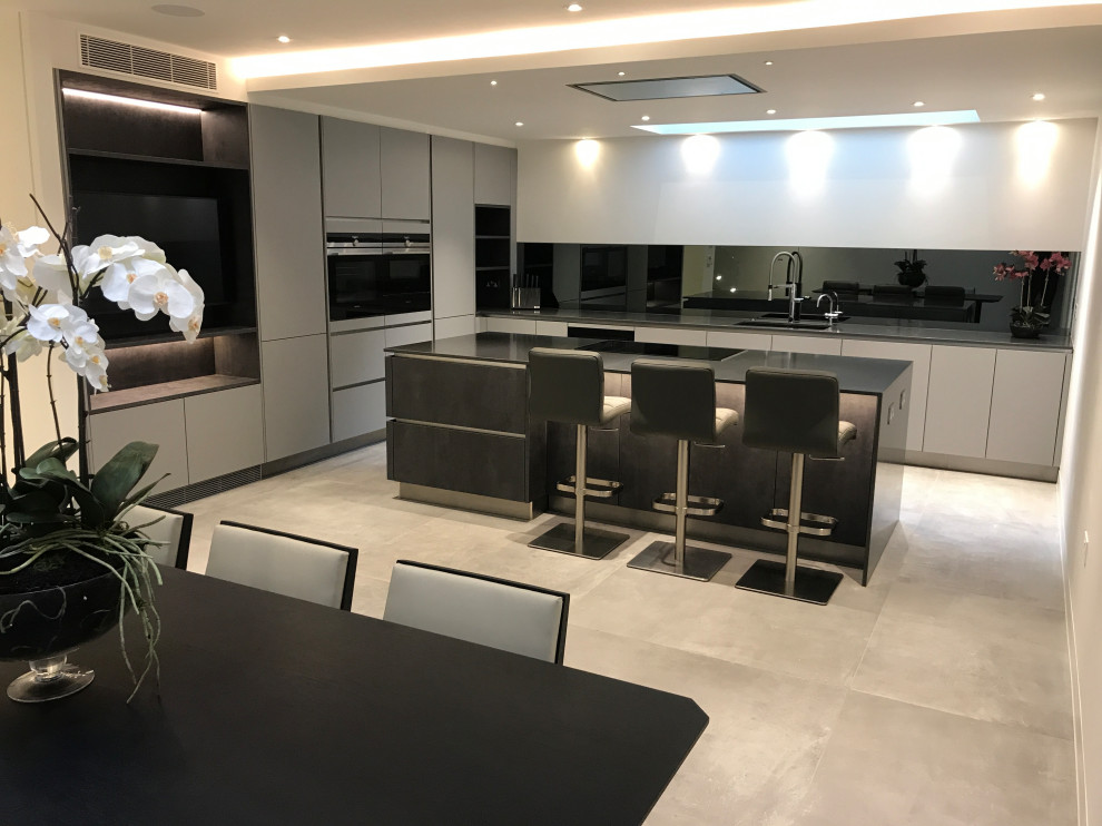 Example of a trendy l-shaped porcelain tile and gray floor eat-in kitchen design in London with a single-bowl sink, flat-panel cabinets, gray cabinets, quartzite countertops, black backsplash, mirror backsplash, black appliances, an island and gray countertops