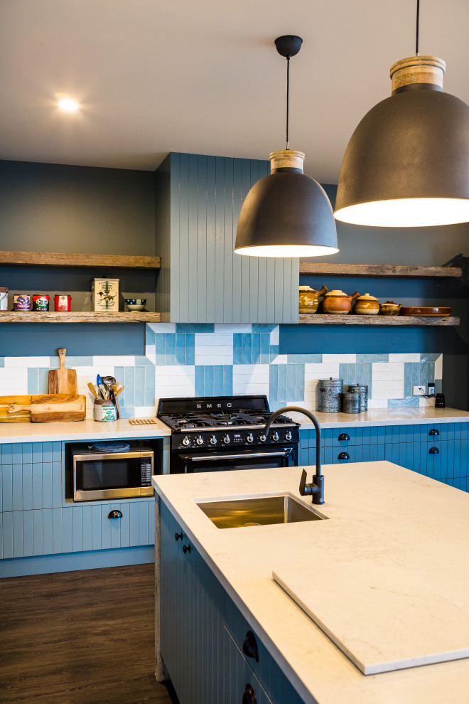 Источник вдохновения для домашнего уюта: угловая кухня-гостиная в современном стиле с накладной мойкой, фасадами с филенкой типа жалюзи, синими фасадами, синим фартуком, фартуком из керамической плитки, черной техникой, темным паркетным полом, островом, коричневым полом и белой столешницей
