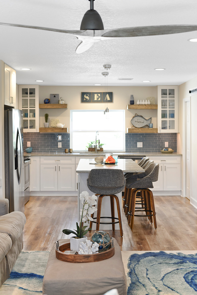 Источник вдохновения для домашнего уюта: угловая кухня-гостиная среднего размера в морском стиле с фасадами в стиле шейкер, белыми фасадами, синим фартуком, фартуком из стеклянной плитки, техникой из нержавеющей стали и островом
