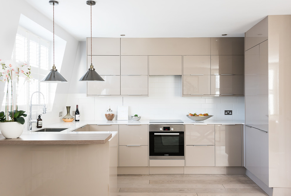 Example of a mid-sized trendy u-shaped light wood floor and beige floor kitchen design in London with an undermount sink, flat-panel cabinets, beige cabinets, white backsplash, subway tile backsplash, paneled appliances and a peninsula