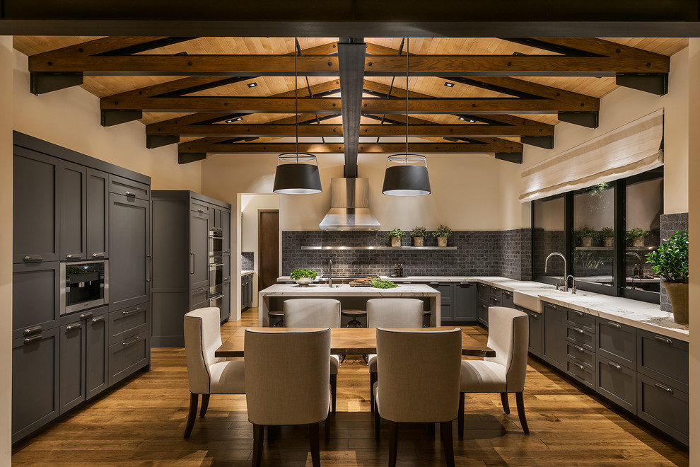 Example of a large trendy l-shaped medium tone wood floor and brown floor eat-in kitchen design in Phoenix with a farmhouse sink, shaker cabinets, gray cabinets, gray backsplash, stainless steel appliances, an island, marble countertops and stone tile backsplash