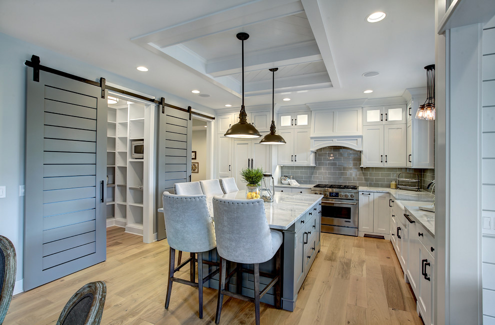 Example of a transitional l-shaped light wood floor and beige floor kitchen design in Grand Rapids with a farmhouse sink, shaker cabinets, white cabinets, marble countertops, gray backsplash, subway tile backsplash, stainless steel appliances and an island