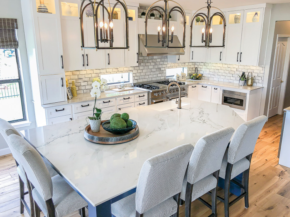 This is an example of a medium sized beach style kitchen/diner in Houston with a belfast sink, recessed-panel cabinets, white cabinets, quartz worktops, grey splashback, ceramic splashback, stainless steel appliances, light hardwood flooring, an island, brown floors and white worktops.