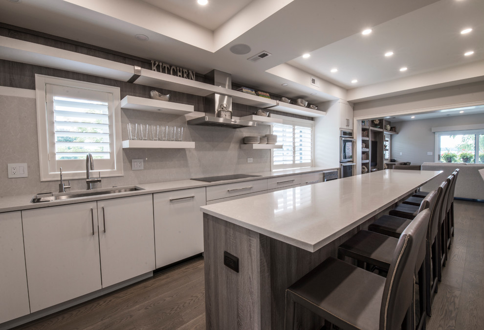 Photo of a medium sized contemporary l-shaped open plan kitchen in Milwaukee with a submerged sink, flat-panel cabinets, distressed cabinets, engineered stone countertops, white splashback, stone slab splashback, integrated appliances, medium hardwood flooring, an island, brown floors and white worktops.
