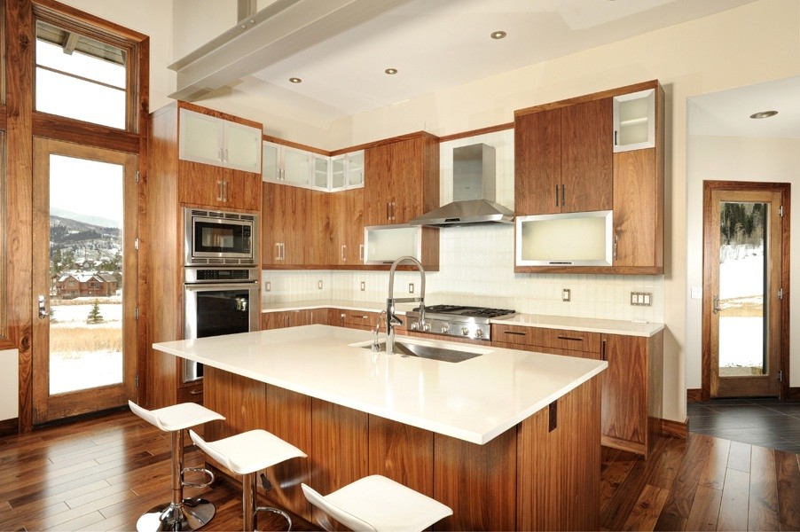 Photo of a medium sized modern l-shaped open plan kitchen in Denver with a submerged sink, flat-panel cabinets, medium wood cabinets, engineered stone countertops, white splashback, glass tiled splashback, stainless steel appliances, medium hardwood flooring, an island and brown floors.