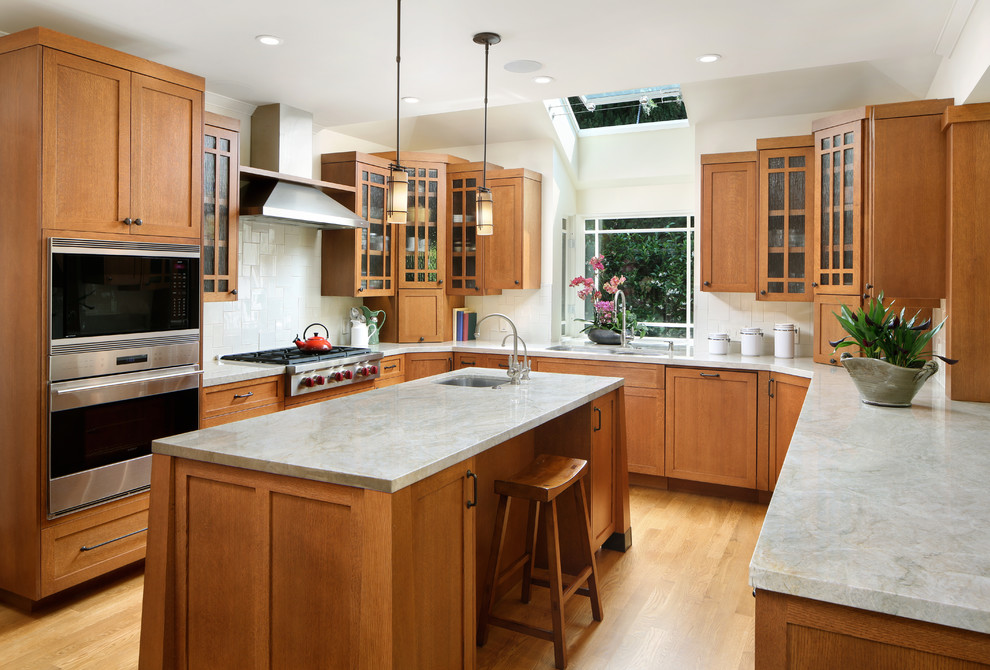 Foto på ett mellanstort amerikanskt u-kök, med en dubbel diskho, skåp i mellenmörkt trä, vitt stänkskydd, ljust trägolv, en köksö, rostfria vitvaror, stänkskydd i keramik, brunt golv och skåp i shakerstil