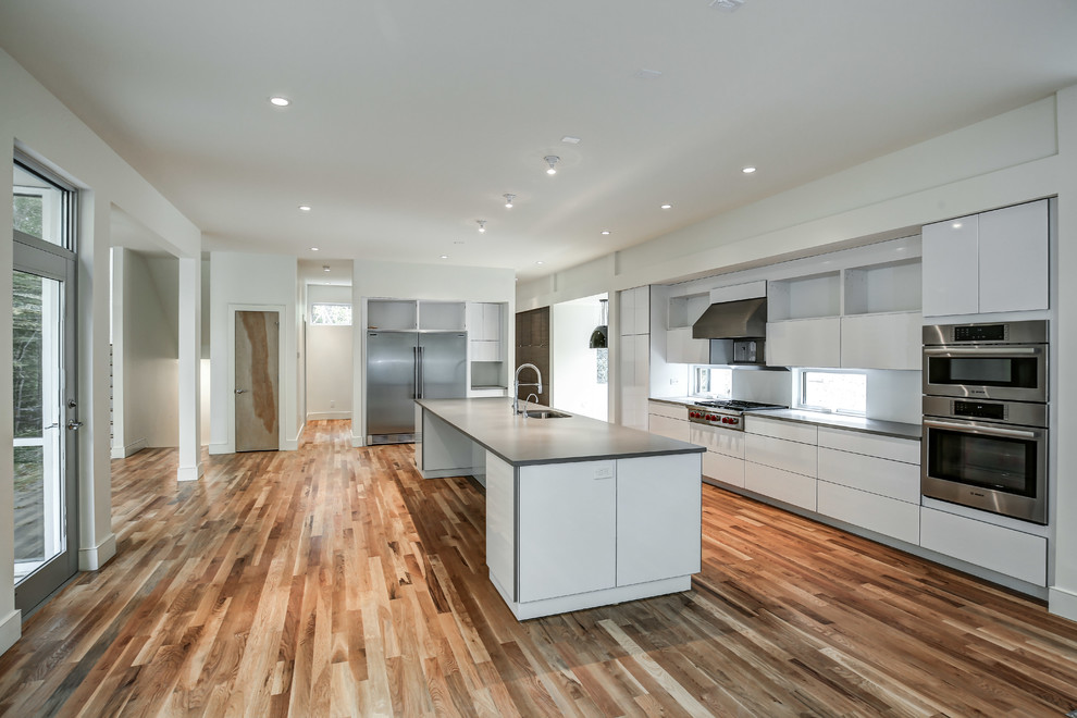 Bild på ett stort funkis l-kök, med en undermonterad diskho, rostfria vitvaror, mellanmörkt trägolv, brunt golv, släta luckor, vita skåp, en köksö och bänkskiva i betong