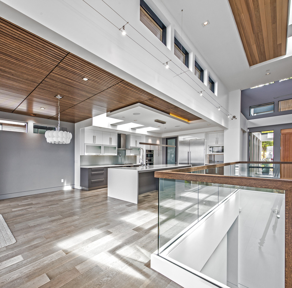Enclosed kitchen - large contemporary l-shaped light wood floor and brown floor enclosed kitchen idea in Seattle with a farmhouse sink, flat-panel cabinets, white cabinets, solid surface countertops, gray backsplash, glass sheet backsplash, stainless steel appliances and an island
