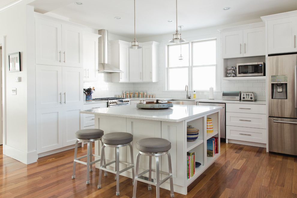 This is an example of a medium sized classic l-shaped open plan kitchen in Boston with a belfast sink, shaker cabinets, white cabinets, white splashback, metro tiled splashback, stainless steel appliances, dark hardwood flooring, an island, brown floors and composite countertops.
