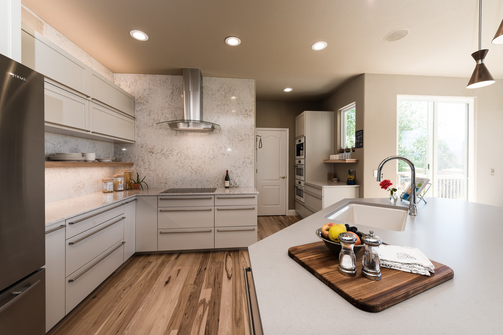 Photo of a medium sized contemporary l-shaped open plan kitchen in Denver with engineered stone countertops, stone slab splashback, stainless steel appliances, an island, a submerged sink, shaker cabinets, white cabinets, white splashback, medium hardwood flooring and brown floors.