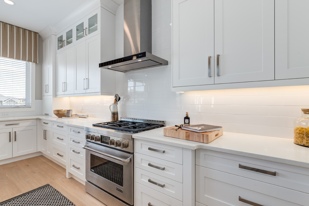 Large contemporary galley kitchen/diner in Calgary with a double-bowl sink, recessed-panel cabinets, white cabinets, engineered stone countertops, white splashback, metro tiled splashback, stainless steel appliances, light hardwood flooring and an island.