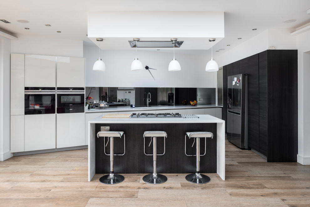 Inspiration for a medium sized contemporary u-shaped kitchen in London with a submerged sink, flat-panel cabinets, mirror splashback, stainless steel appliances, light hardwood flooring, an island, beige floors, white worktops and black cabinets.