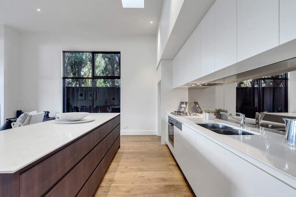 Inspiration for a large contemporary single-wall open plan kitchen in Adelaide with a submerged sink, flat-panel cabinets, white cabinets, engineered stone countertops, metallic splashback, mirror splashback, medium hardwood flooring, an island, white worktops and brown floors.