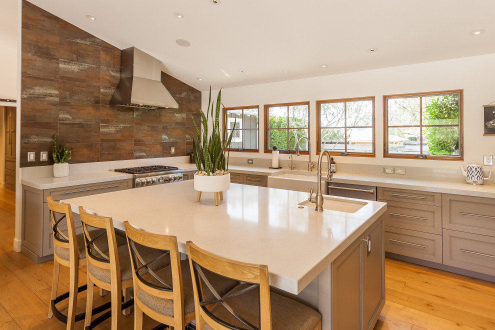 Example of a country u-shaped medium tone wood floor, brown floor and vaulted ceiling kitchen design in San Luis Obispo with a farmhouse sink, recessed-panel cabinets, gray cabinets, brown backsplash, stainless steel appliances, an island and white countertops