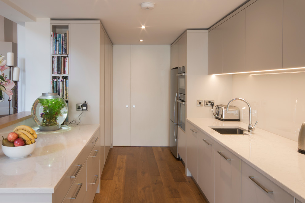 Example of a mid-sized trendy galley medium tone wood floor enclosed kitchen design in Other with a drop-in sink, flat-panel cabinets, beige cabinets, quartz countertops, white backsplash, glass sheet backsplash and stainless steel appliances