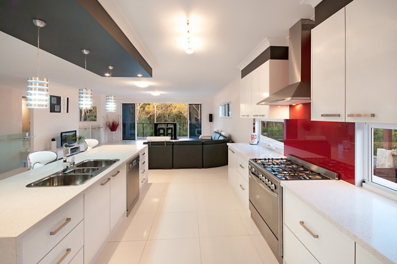 Foto på ett stort funkis vit kök, med en trippel diskho, vita skåp, bänkskiva i kvarts, rött stänkskydd, glaspanel som stänkskydd, rostfria vitvaror, klinkergolv i porslin, en köksö och vitt golv