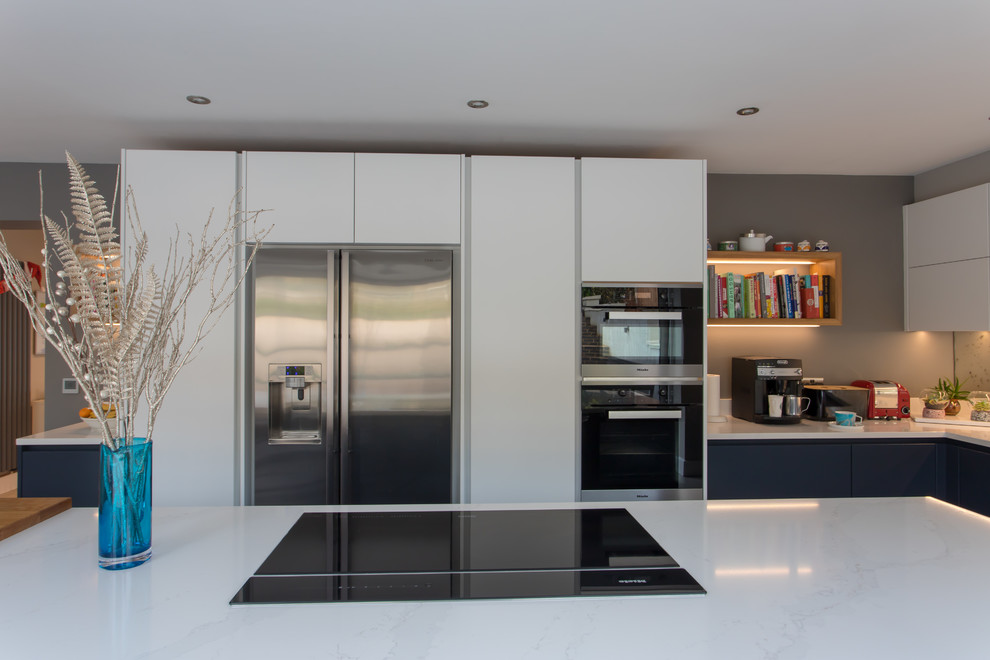 This is an example of an expansive contemporary l-shaped kitchen/diner in Hertfordshire with a built-in sink, flat-panel cabinets, blue cabinets, quartz worktops, metallic splashback, mirror splashback, stainless steel appliances, porcelain flooring, an island, white floors and white worktops.