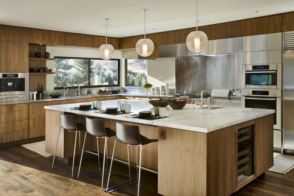 Foto på ett funkis l-kök, med släta luckor, skåp i mellenmörkt trä, bänkskiva i rostfritt stål, stänkskydd med metallisk yta, rostfria vitvaror, mörkt trägolv och en köksö
