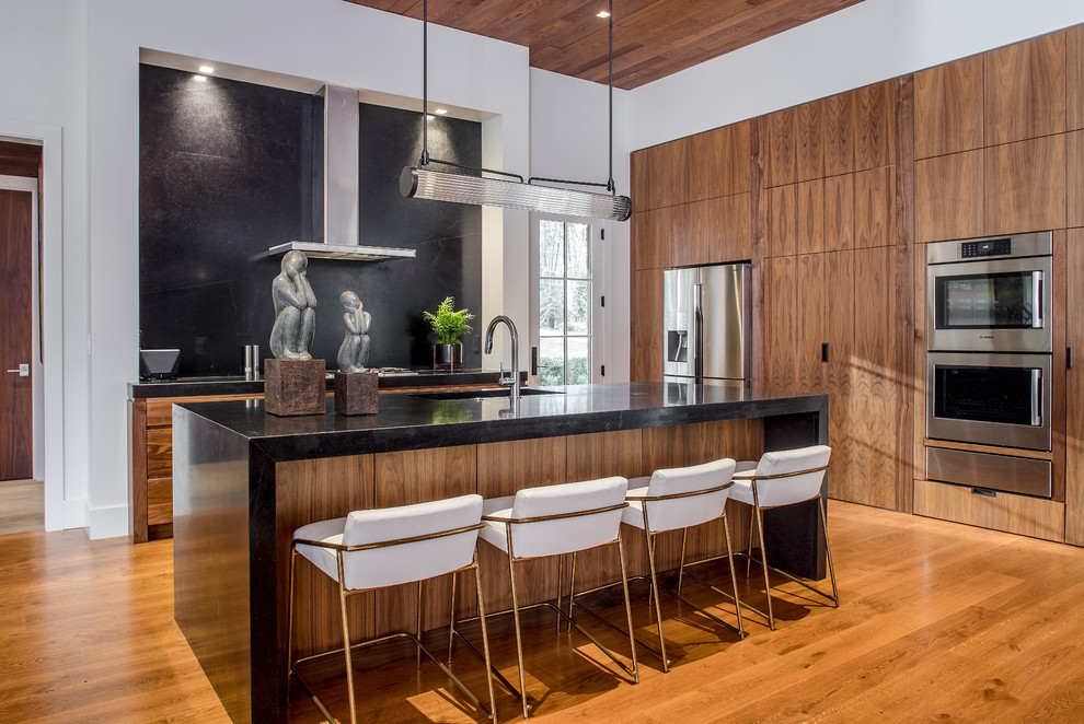 Cette image montre une cuisine design en L et bois brun avec un évier encastré, un placard à porte plane, un électroménager en acier inoxydable, un sol en bois brun, îlot, un sol marron et plan de travail noir.