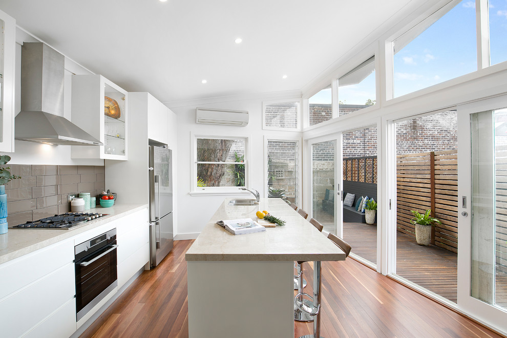 Photo of a contemporary galley kitchen in Sydney with a double-bowl sink, flat-panel cabinets, white cabinets, marble worktops, brown splashback, stainless steel appliances, medium hardwood flooring, an island, brown floors and beige worktops.