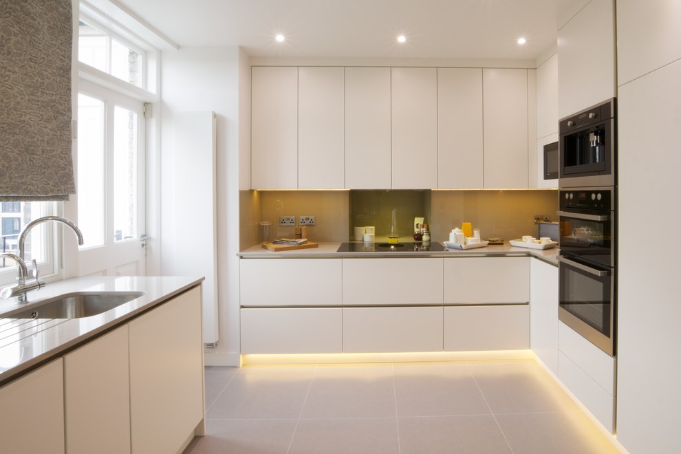 Example of a transitional u-shaped beige floor kitchen design in London with an undermount sink, flat-panel cabinets, white cabinets, beige backsplash, stainless steel appliances and beige countertops
