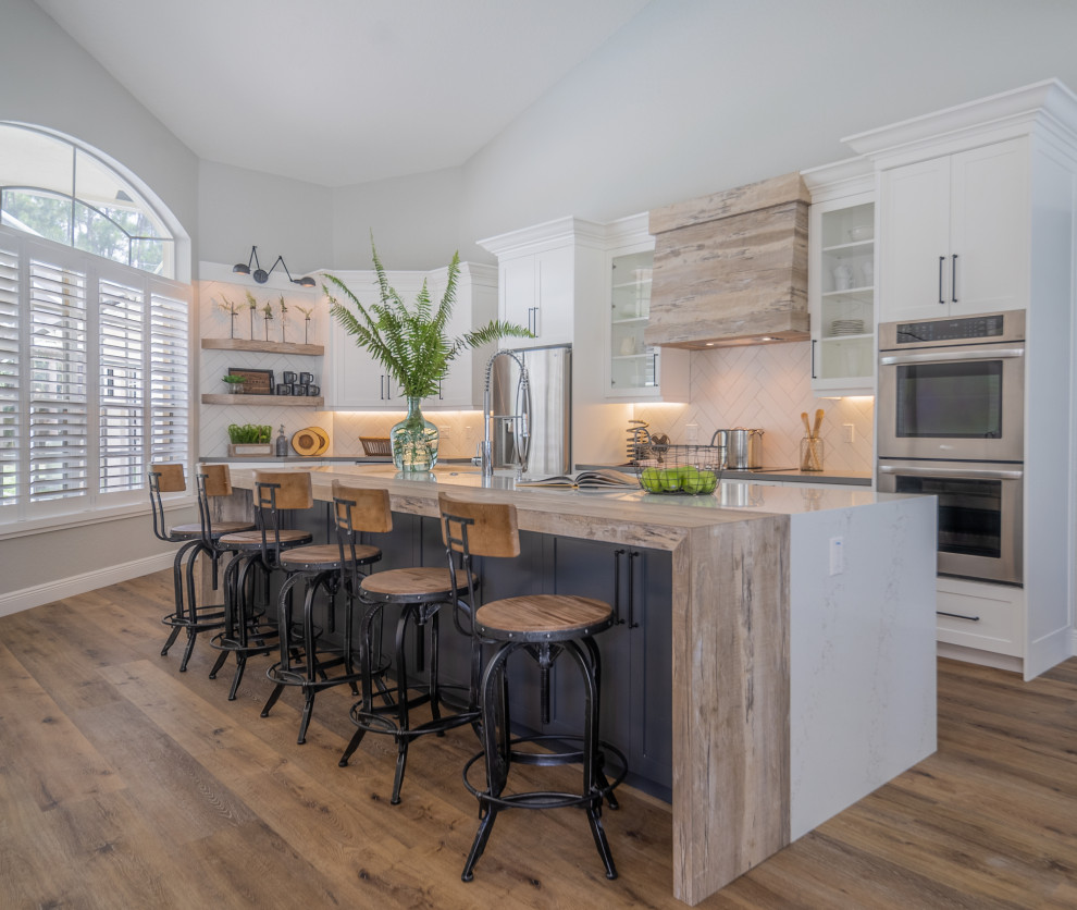 Photo of a large classic l-shaped kitchen with an integrated sink, shaker cabinets, white cabinets, quartz worktops, white splashback, porcelain splashback, stainless steel appliances, vinyl flooring, an island, brown floors and white worktops.