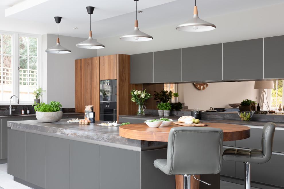 Large contemporary open plan kitchen in Hampshire with an integrated sink, glass-front cabinets, grey cabinets, quartz worktops, metallic splashback, mirror splashback, black appliances, an island and grey worktops.