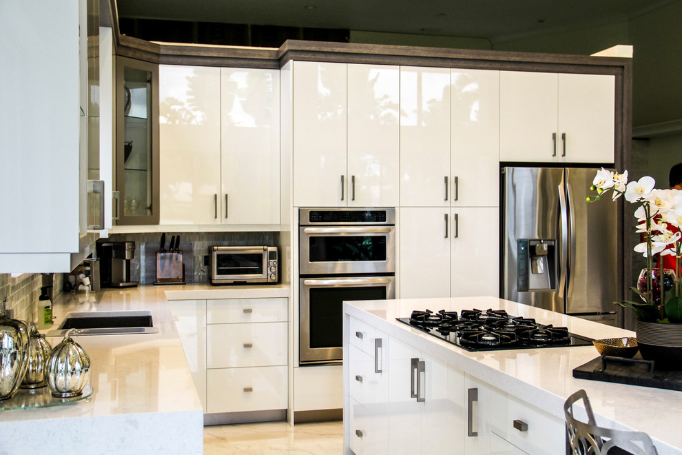 This is an example of a medium sized contemporary l-shaped kitchen/diner in Miami with glass-front cabinets, white cabinets, grey splashback, a submerged sink, engineered stone countertops, glass tiled splashback, stainless steel appliances, marble flooring, an island and beige floors.