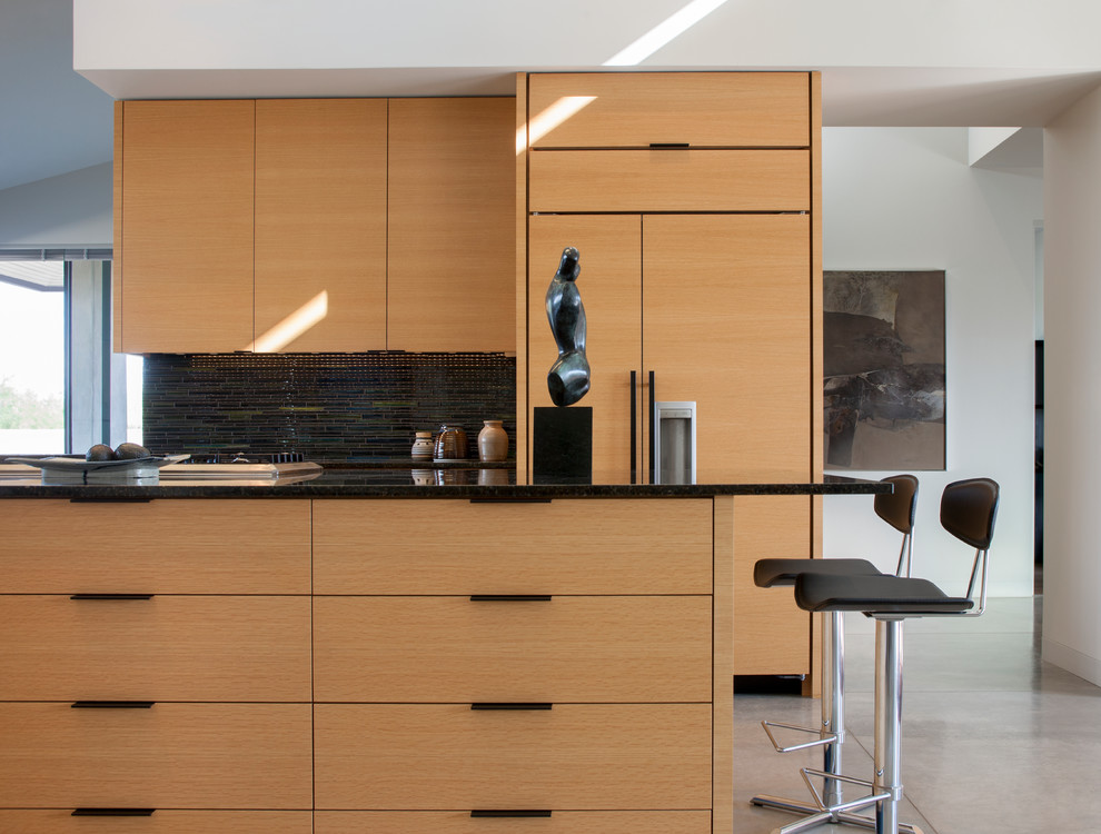 Moderne Küche mit flächenbündigen Schrankfronten, hellbraunen Holzschränken, Küchenrückwand in Schwarz, Kücheninsel und grauem Boden in Santa Barbara