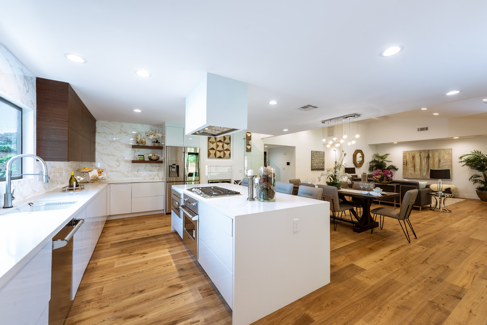 Inspiration for a large contemporary l-shaped open plan kitchen in Los Angeles with a submerged sink, flat-panel cabinets, white cabinets, engineered stone countertops, white splashback, porcelain splashback, stainless steel appliances, light hardwood flooring, an island, white worktops and brown floors.