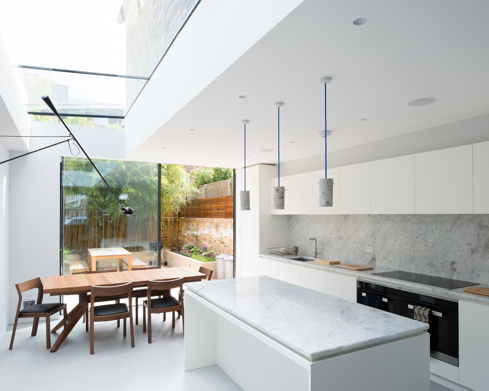Contemporary galley kitchen/diner in London with a submerged sink, flat-panel cabinets, white cabinets, white splashback, stone slab splashback, black appliances and an island.