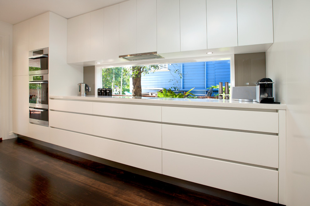 Photo of a large contemporary galley kitchen/diner in Brisbane with a double-bowl sink, flat-panel cabinets, white cabinets, engineered stone countertops, brown splashback, glass tiled splashback, stainless steel appliances, an island and dark hardwood flooring.