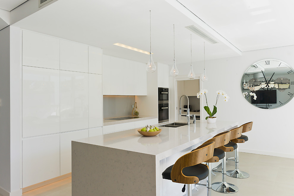 На фото: параллельная кухня в современном стиле с врезной мойкой, белыми фасадами, серым фартуком, островом, черной техникой и полом из керамогранита с