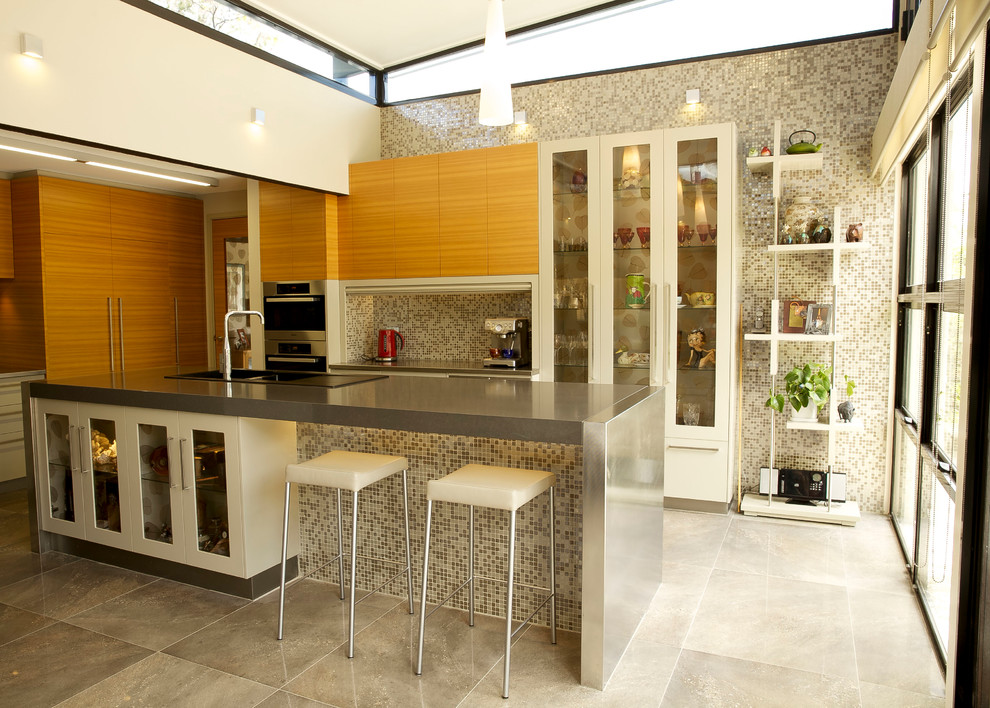 Modern inredning av ett u-kök, med en nedsänkt diskho, släta luckor, skåp i ljust trä, bänkskiva i rostfritt stål, grått stänkskydd, stänkskydd i mosaik, integrerade vitvaror och en köksö