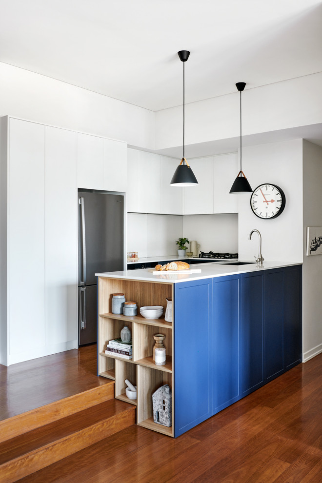 Bild på ett mellanstort funkis vit vitt kök, med en undermonterad diskho, skåp i shakerstil, blå skåp, bänkskiva i kvarts, vitt stänkskydd, stänkskydd i sten, rostfria vitvaror, mellanmörkt trägolv, en halv köksö och brunt golv