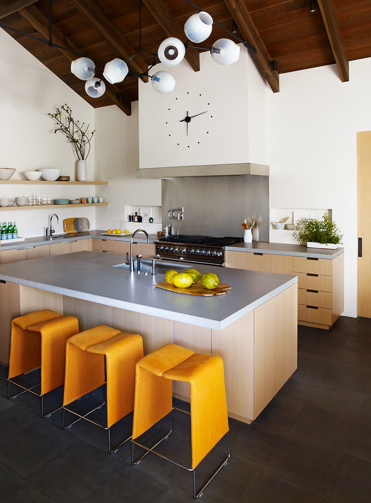Modern inredning av ett l-kök, med en integrerad diskho, släta luckor, skåp i ljust trä, bänkskiva i betong, rostfria vitvaror, en köksö och svart golv