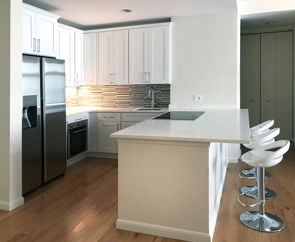 Small trendy u-shaped light wood floor and brown floor eat-in kitchen photo in New York with a drop-in sink, shaker cabinets, white cabinets, quartz countertops, metallic backsplash, stainless steel appliances and a peninsula