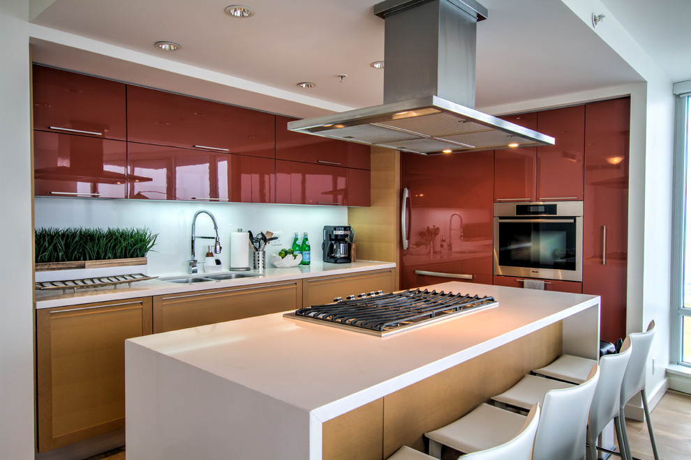 Foto di un cucina con isola centrale minimal con lavello a doppia vasca, ante lisce, ante rosse, paraspruzzi con lastra di vetro e elettrodomestici in acciaio inossidabile