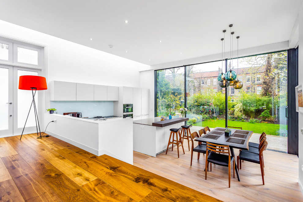 Photo of a contemporary galley kitchen/diner in London with flat-panel cabinets, white cabinets, glass sheet splashback, stainless steel appliances, light hardwood flooring and an island.