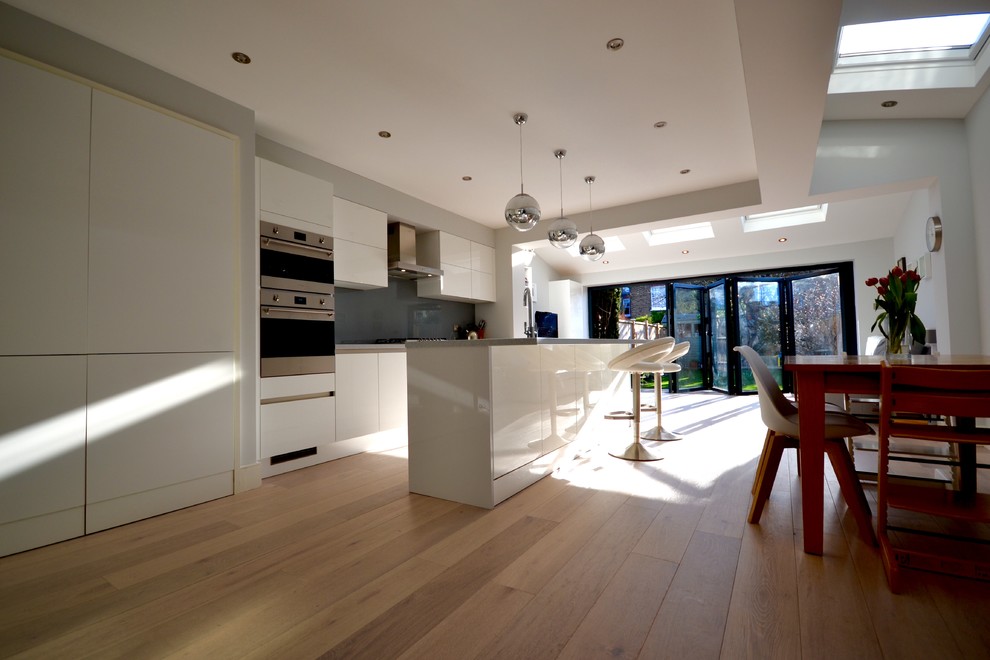 Photo of a medium sized contemporary single-wall open plan kitchen in London with an integrated sink, flat-panel cabinets, grey cabinets, composite countertops, grey splashback, glass sheet splashback, stainless steel appliances, medium hardwood flooring and a breakfast bar.