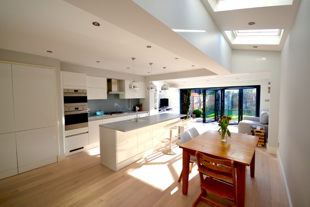 Medium sized contemporary single-wall open plan kitchen in London with an integrated sink, flat-panel cabinets, grey cabinets, composite countertops, grey splashback, glass sheet splashback, stainless steel appliances, medium hardwood flooring and a breakfast bar.