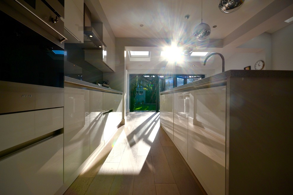 This is an example of a medium sized contemporary single-wall open plan kitchen in London with an integrated sink, flat-panel cabinets, grey cabinets, composite countertops, grey splashback, glass sheet splashback, stainless steel appliances, medium hardwood flooring and a breakfast bar.