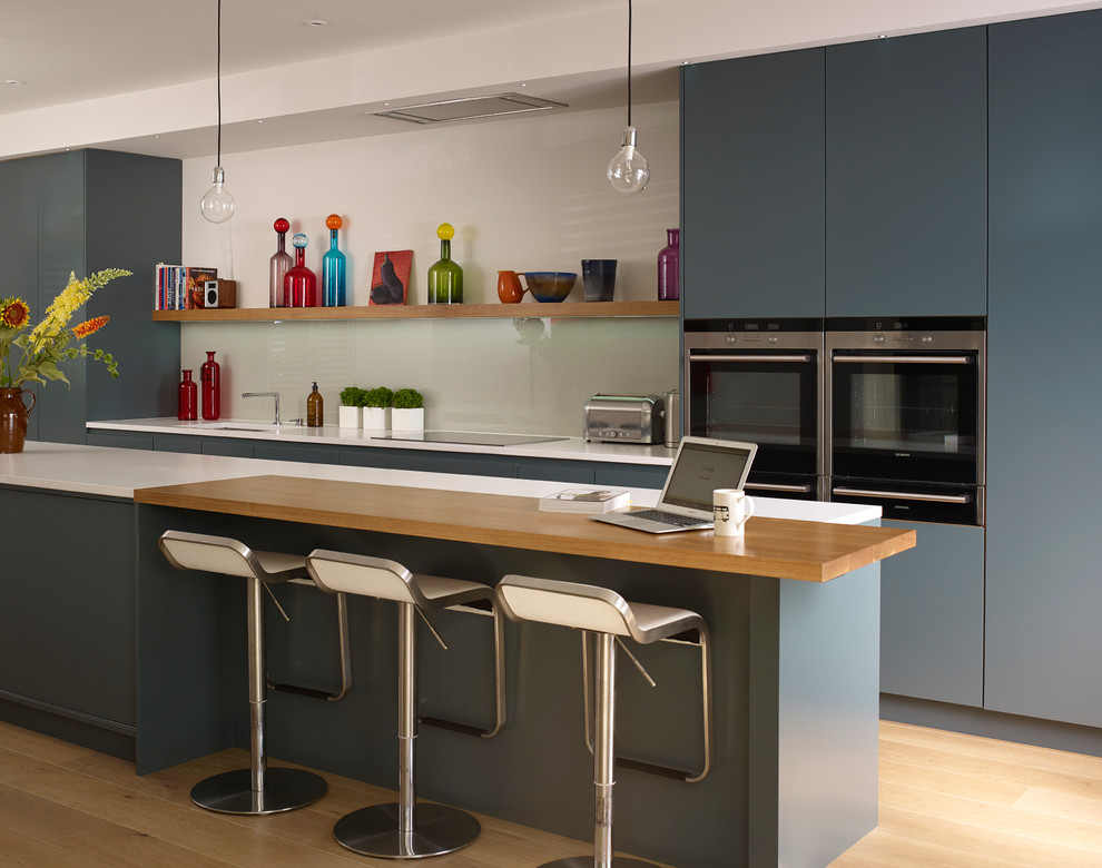 Modelo de cocina contemporánea con armarios con paneles lisos, puertas de armario azules, salpicadero blanco, electrodomésticos de acero inoxidable, suelo de madera clara, una isla y barras de cocina
