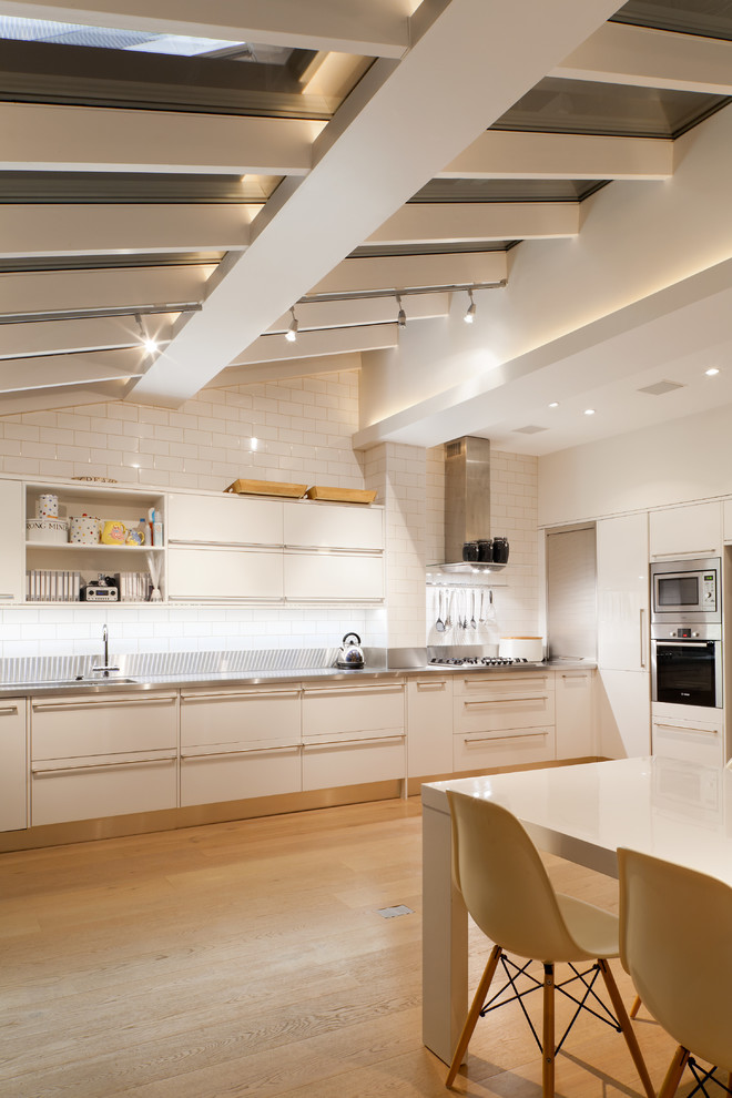 Moderne Wohnküche ohne Insel mit weißen Schränken, flächenbündigen Schrankfronten, Küchenrückwand in Weiß, Rückwand aus Metrofliesen, Küchengeräten aus Edelstahl und hellem Holzboden in London