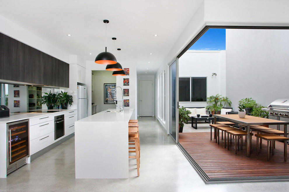 На фото: параллельная кухня-гостиная в современном стиле с плоскими фасадами, белыми фасадами, зеркальным фартуком, бетонным полом, островом и техникой из нержавеющей стали с