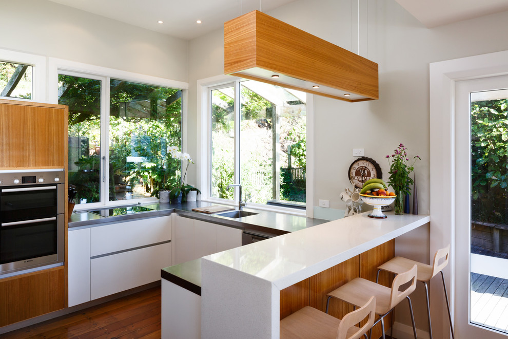 Moderne Küche mit integriertem Waschbecken, flächenbündigen Schrankfronten, weißen Schränken, Küchengeräten aus Edelstahl, braunem Holzboden und Halbinsel in Wellington