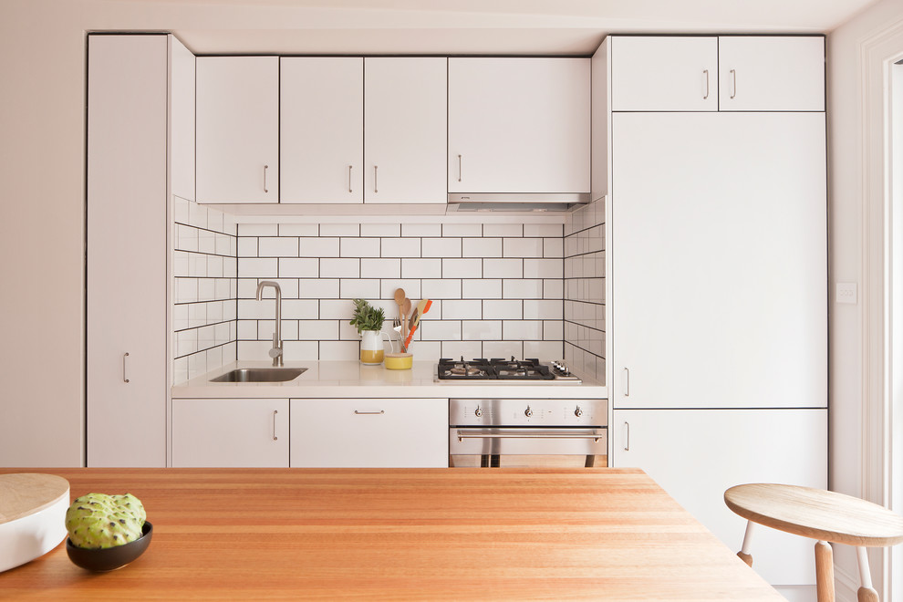 Bild på ett funkis linjärt kök, med en undermonterad diskho, släta luckor, vita skåp, träbänkskiva, vitt stänkskydd, stänkskydd i tunnelbanekakel, rostfria vitvaror och en köksö