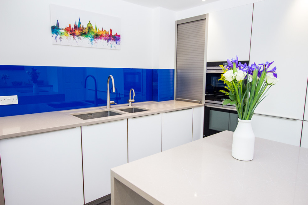 オックスフォードシャーにある高級な広いコンテンポラリースタイルのおしゃれなアイランドキッチン (一体型シンク、フラットパネル扉のキャビネット、白いキャビネット、珪岩カウンター、青いキッチンパネル、ガラスタイルのキッチンパネル、シルバーの調理設備、無垢フローリング、グレーの床) の写真