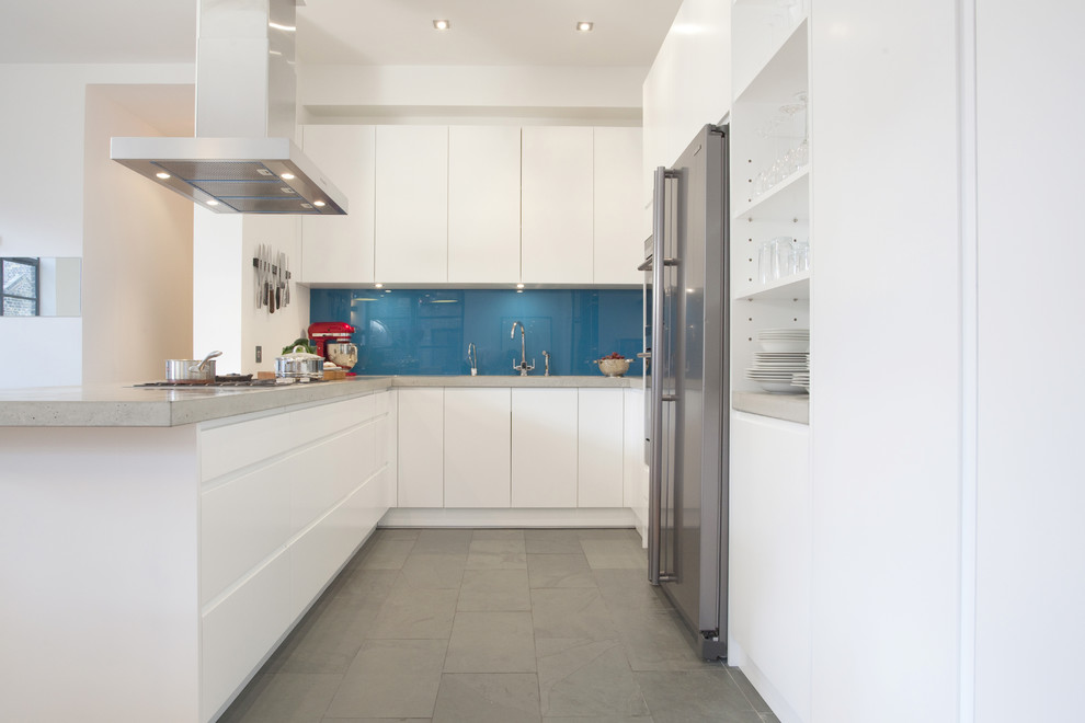 Trendy u-shaped kitchen photo in London with stainless steel appliances, concrete countertops, flat-panel cabinets, white cabinets, blue backsplash and glass sheet backsplash
