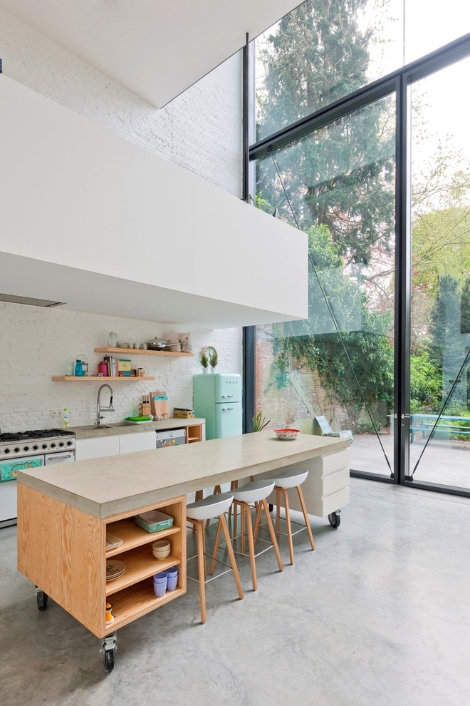 Foto på ett stort funkis linjärt kök och matrum, med en integrerad diskho, släta luckor, vita skåp, bänkskiva i betong, vitt stänkskydd, färgglada vitvaror, betonggolv och en köksö
